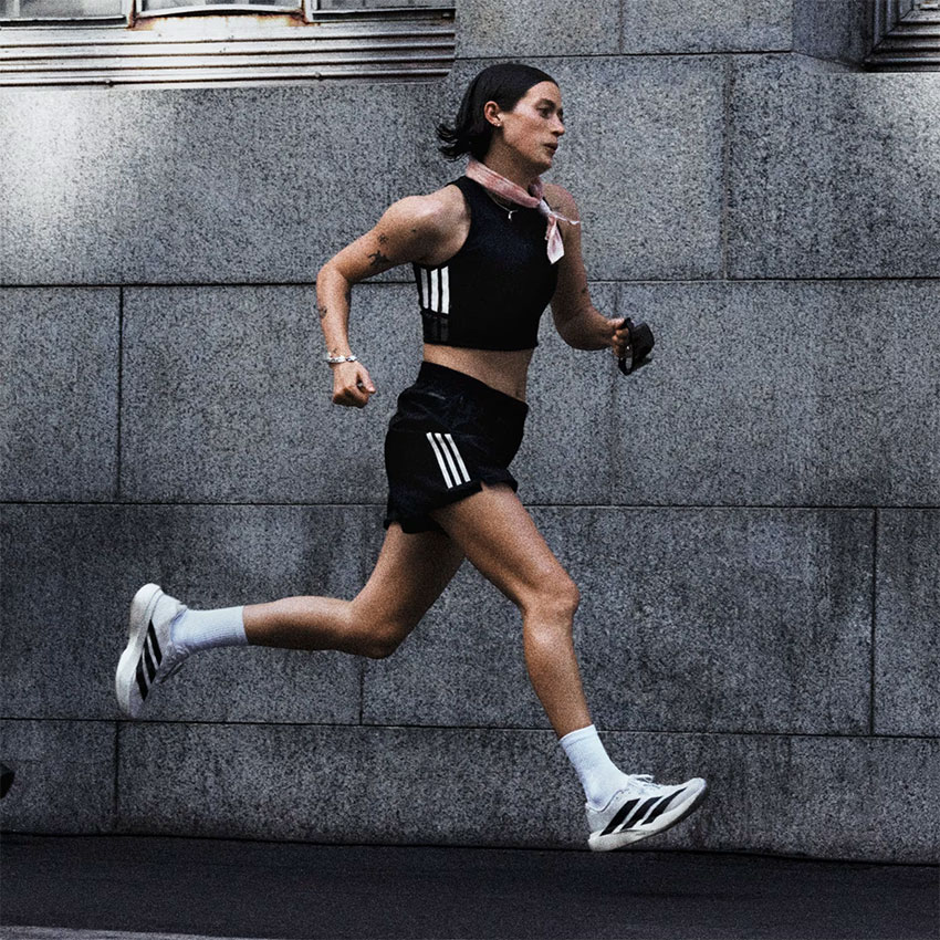 Wanita berlari menggunakan Running Shoes Wanita Terbaik 2025 warna putih dengan garis hitam, dipadukan dengan pakaian olahraga hitam dan kaos kaki putih, di depan dinding beton — sumber foto www.adidas.co.za