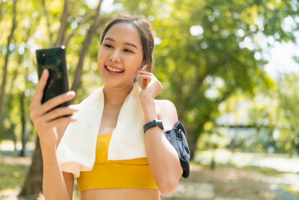 Wanita sedang tersenyum sambil melihat ponsel setelah berlari, mengenakan sport bra kuning dan handuk di leher, menggambarkan konsep Menghasilkan Uang dari Kegiatan Running melalui smartphone – Sumber foto: Freepik.