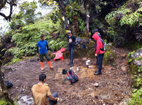 Pendaki sedang melakukan manajemen istirahat di gunung, berhenti di area datar untuk mengatur tenaga, minum, dan memeriksa perlengkapan sebelum melanjutkan perjalanan mendaki di jalur hutan basah.