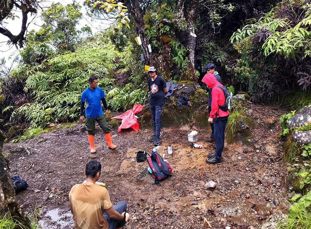 Pendaki sedang melakukan manajemen istirahat di gunung, berhenti di area datar untuk mengatur tenaga, minum, dan memeriksa perlengkapan sebelum melanjutkan perjalanan mendaki di jalur hutan basah.