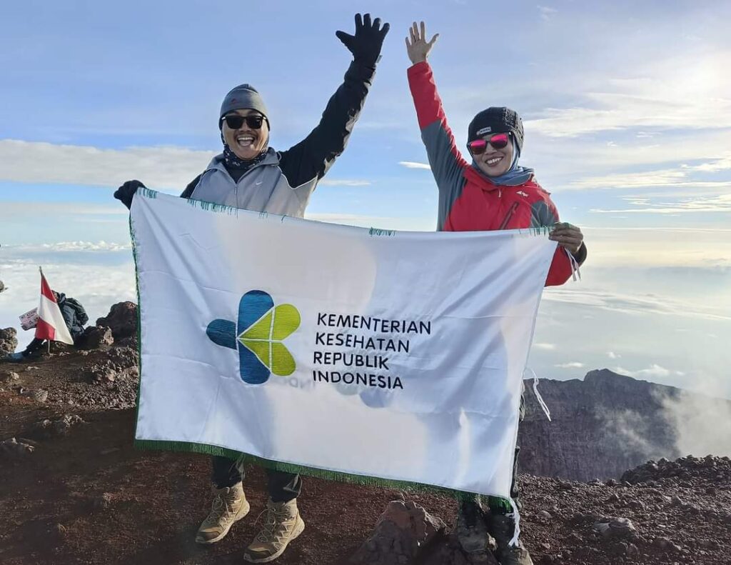 Dua pendaki dari Kementerian Kesehatan Republik Indonesia berfoto di puncak Gunung Kerinci sambil membentangkan bendera, dengan latar awan dan langit cerah. Foto ini diambil dalam pendakian bersama trip operator Kerinci Earth. (Sumber Foto: Instagram @kerinciearth